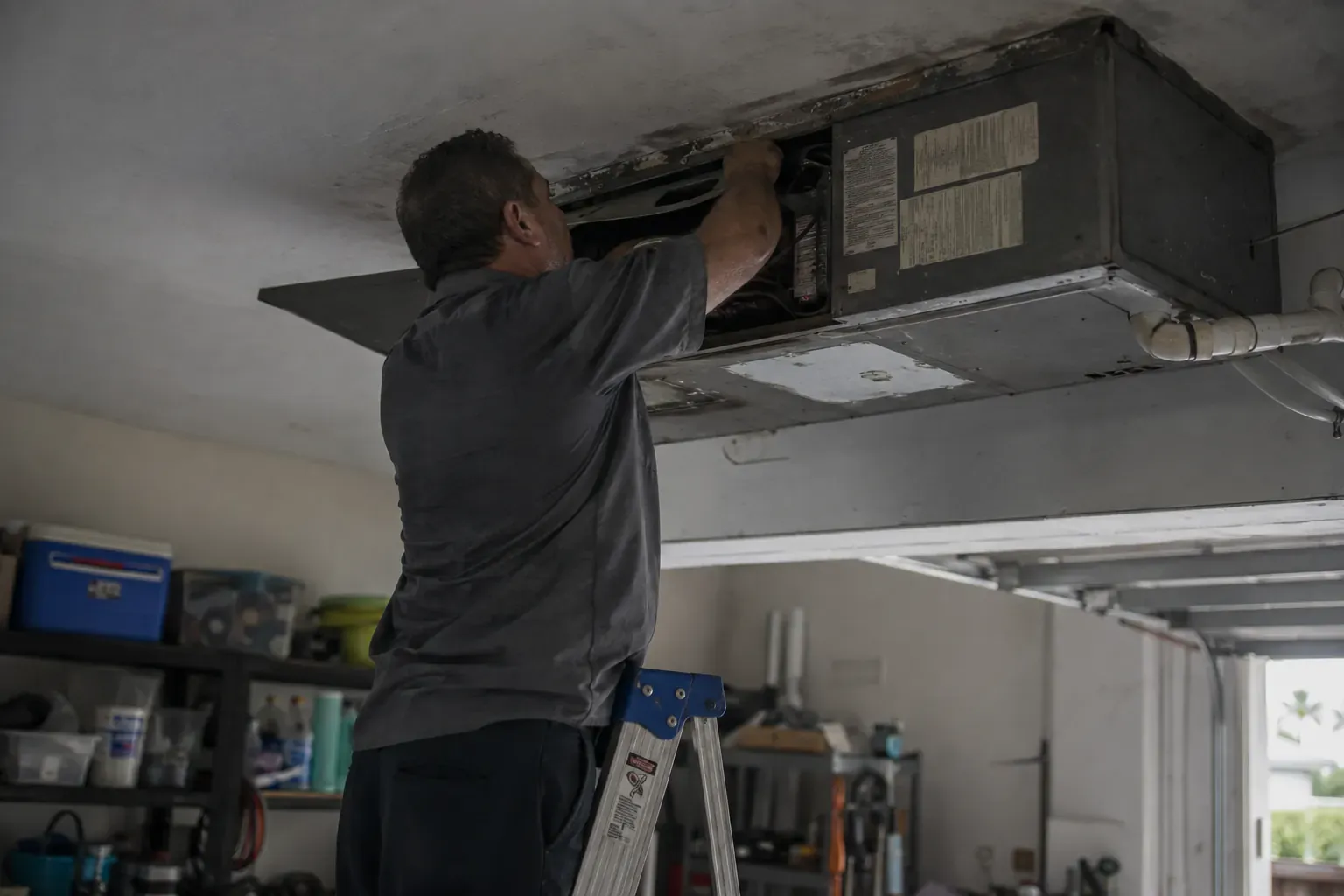 Technician on a step ladder inspecting a ceiling-mounted air handler in a garage in Gateway, FL