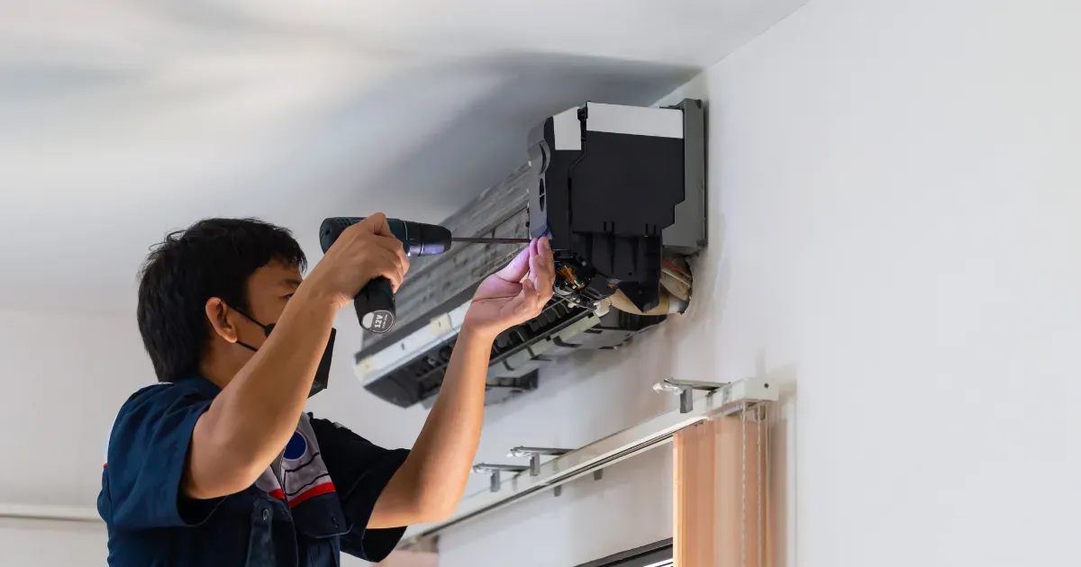 A technician uses a power drill to remove the front panel of a wall-mounted air conditioning unit during a repair or maintenance service