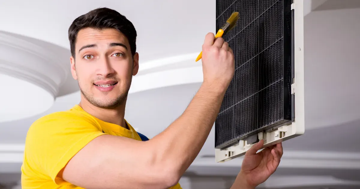 Technician in a yellow shirt cleaning an air conditioning filter with a small brush in a bright indoor space