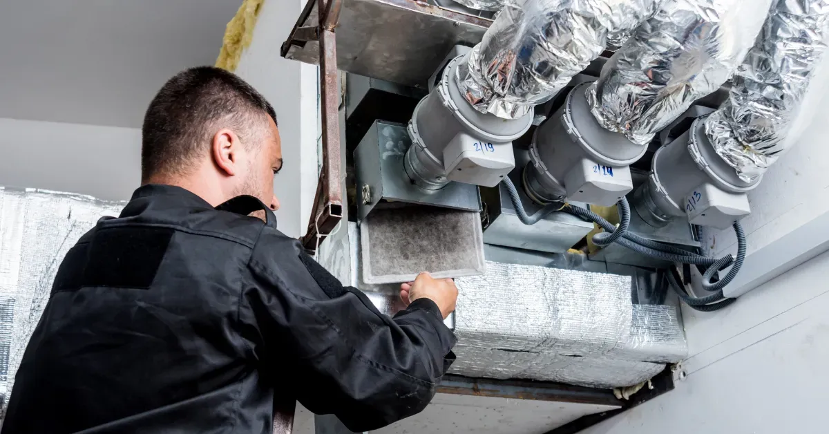 A technician removing a dirty air filter from a duct system, surrounded by insulated ventilation pipes
